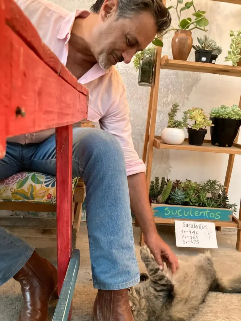 Omar Germenos, copropietario de GP Botas Artesanales, usando botas de cuero café hechas a mano mientras acaricia a un gato junto a un estante de madera con suculentas.